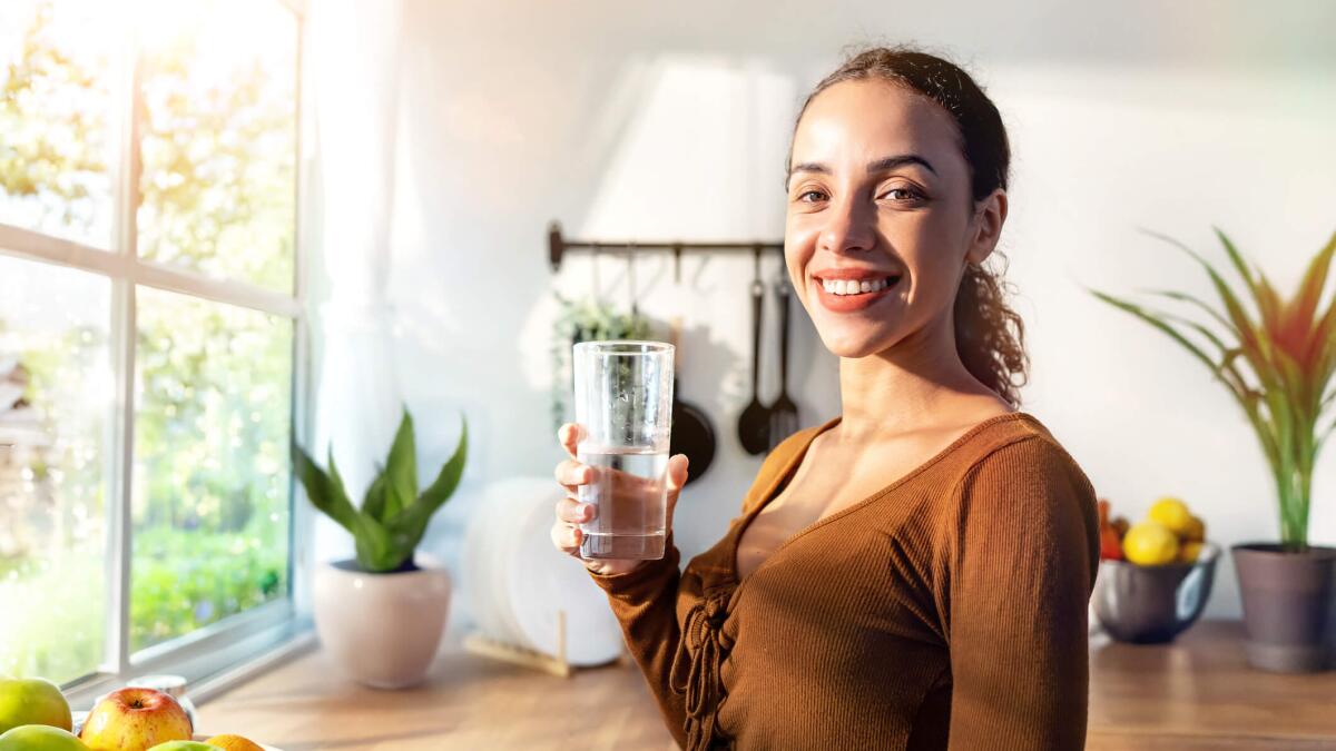 Eine lächelnde Frau steht in einer Küche und hat ein Glas Wasser in der Hand. Durch das Fenster scheint die Sonne auf sie