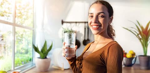 Eine lächelnde Frau steht in einer Küche und hat ein Glas Wasser in der Hand. Durch das Fenster scheint die Sonne auf sie