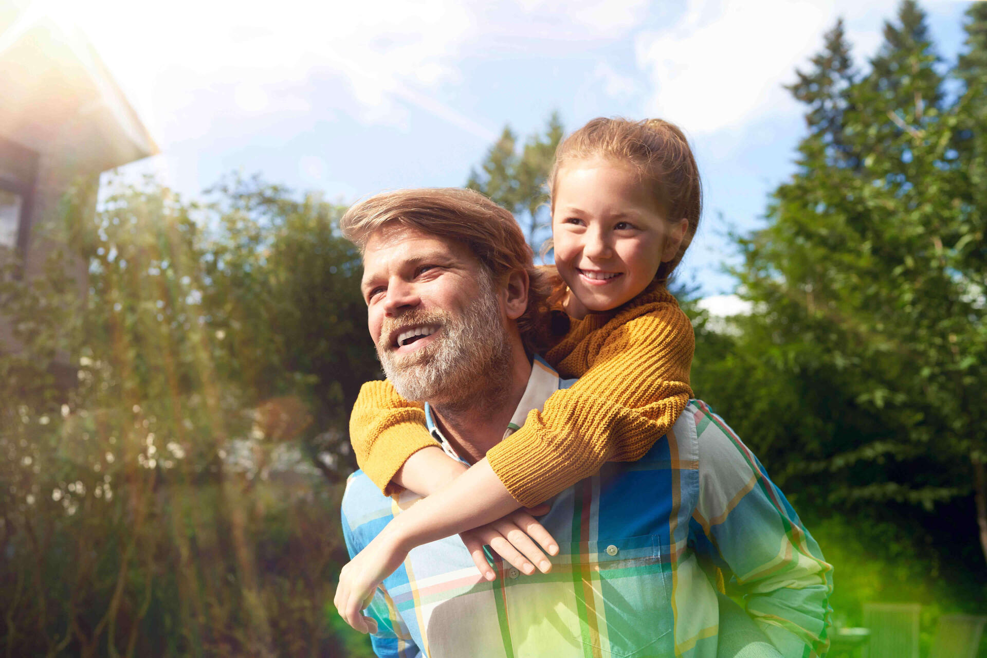 Ein Vater trägt seine Tochter auf dem Rücken. Beide lächeln und die Sonne scheint