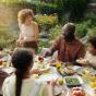 Eine Gruppe Menschen sitzt in einem schönen Garten an einem gedeckte Tisch und isst gemeinsam