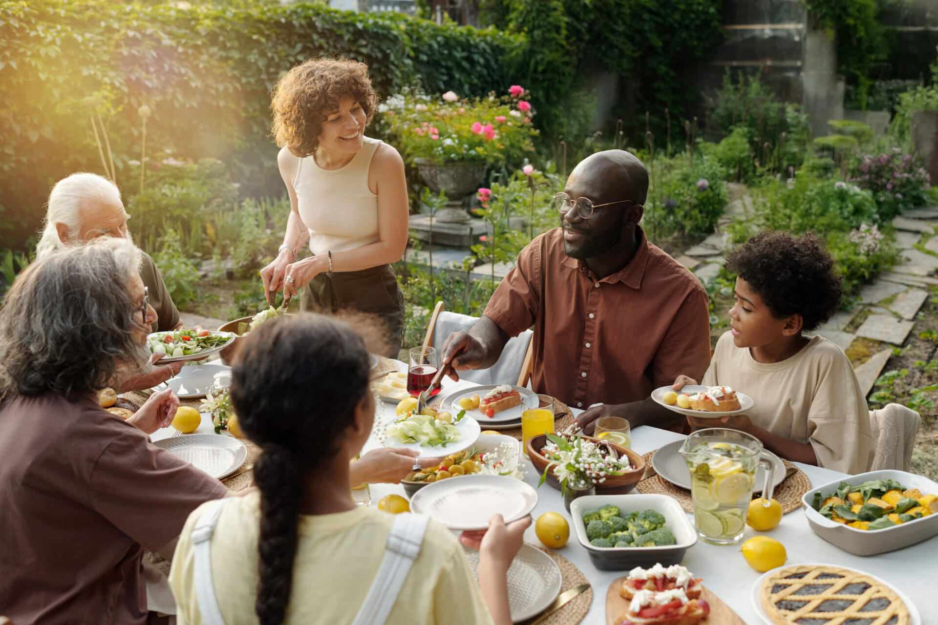 Eine Gruppe Menschen sitzt in einem schönen Garten an einem gedeckte Tisch und isst gemeinsam