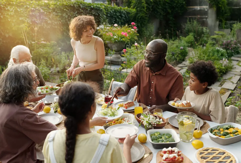Eine Gruppe Menschen sitzt in einem schönen Garten an einem gedeckte Tisch und isst gemeinsam