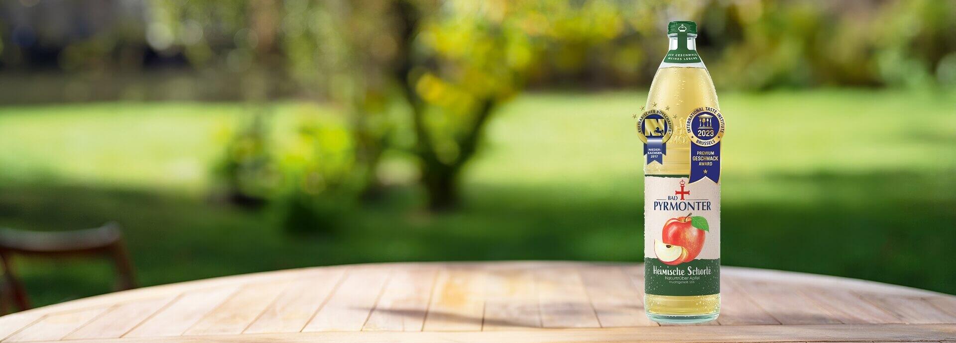Eine Glasflasche Bad Pyrmonter Heimische Schorle naturtrüber Apfel auf einem Tisch vor einem Naturhintergrund mit mehreren Auszeichnungen