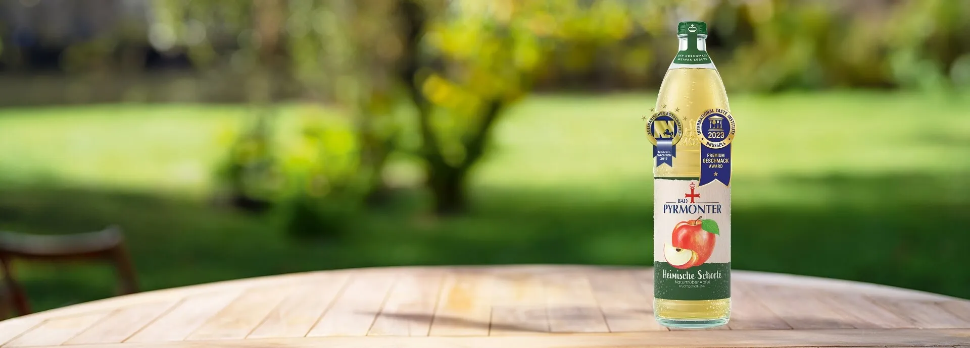 Eine Glasflasche Bad Pyrmonter Heimische Schorle naturtrüber Apfel auf einem Tisch vor einem Naturhintergrund mit mehreren Auszeichnungen